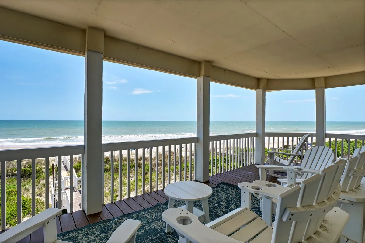 The image captures a stunning oceanfront deck with Adirondack chairs overlooking the serene waves of North Topsail Beach. The spacious deck is bathed in natural light, inviting guests to relax, gather, and take in panoramic ocean views. Subtle details evoke the possibilities of dolphin watching, enjoying the salty breeze, or unwinding beneath the stars.