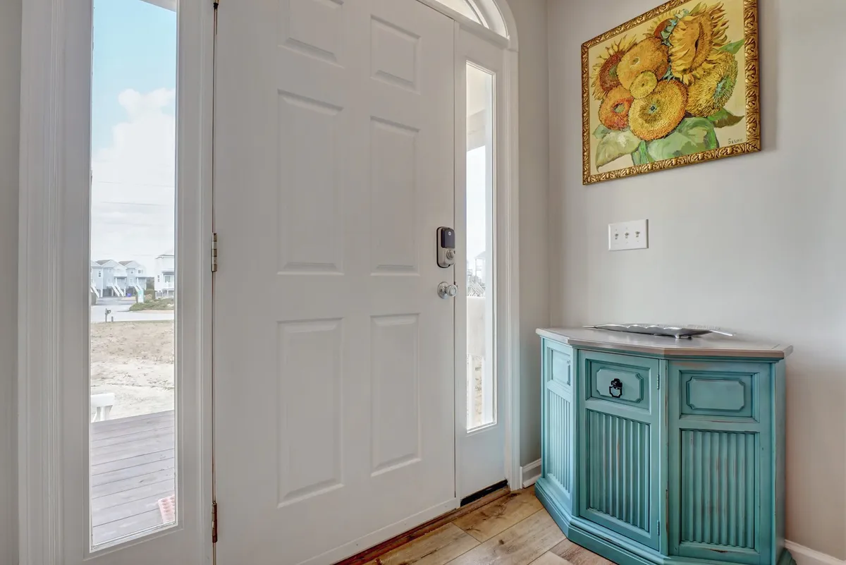 A bright and inviting entryway showcasing a blue console table, a cheerful sunflower painting, and the front door welcoming guests into the home. The space is filled with light, setting an organized and positive first impression.
