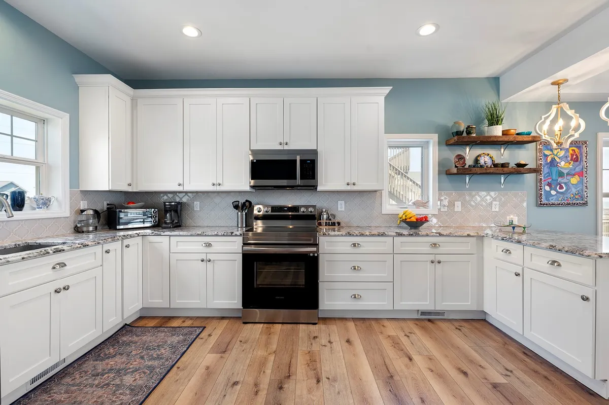 A spacious kitchen with stainless steel appliances, granite countertops, open shelving adorned with decorative items, and a large window providing plenty of natural light. The scene suggests a welcoming, communal area designed for family gatherings and personal touches.