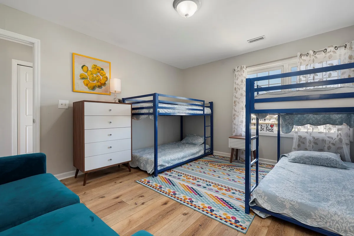 A cheerful bedroom with bunk beds, a colorful area rug, a chest of drawers, and a window with curtains. The space exudes a playful, family-friendly vibe suitable for kids or groups.