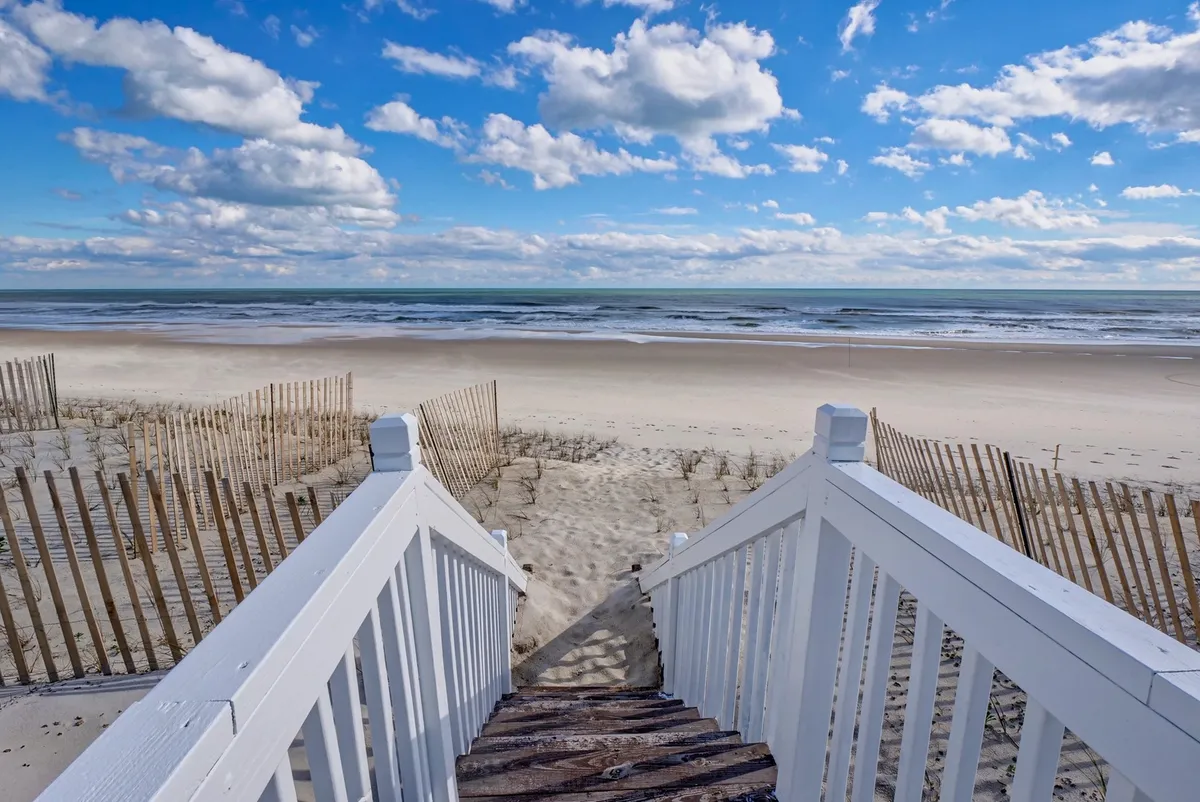 A wide staircase leads directly onto the sandy beach, with ocean waves rolling in and clouds scattered across the sky. The scene evokes outdoor fun and easy access to nature, ideal for all ages of the family.