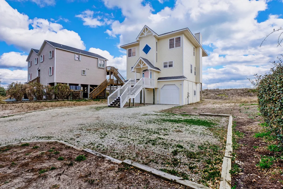 A welcoming exterior shot of the large, three-story beach house with an inviting staircase and a unique diamond-shaped window. The house is set against a pleasant outdoor backdrop, perfect for family gatherings and post-beach relaxation.