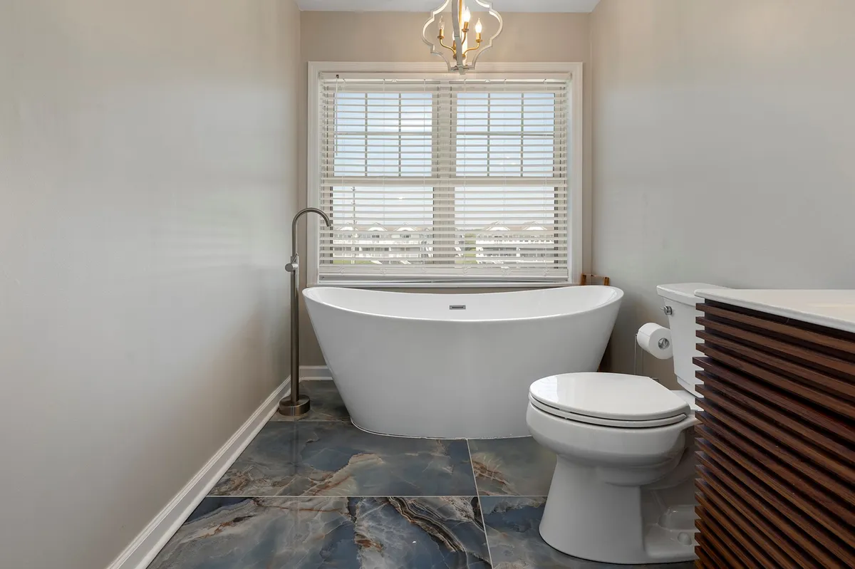 A stylish bathroom interior featuring a freestanding bathtub, a window with blinds, an elegant chandelier, and textured flooring. The mood is tranquil and spa-like, with natural light inviting relaxation and self-care.