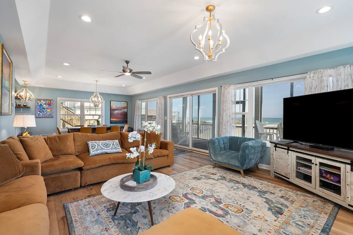 This image showcases a comfortable brown sofa arranged on a colorful area rug, with large windows revealing a sweeping ocean view. The modern chandelier above and the TV unit with tasteful decor complete the inviting atmosphere. Sunlight streams in, illuminating the space and creating a cozy yet open environment perfect for relaxing or gathering.