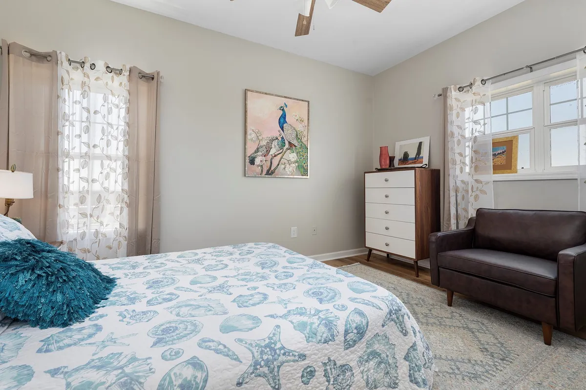 A serene bedroom featuring a bed with sea-themed bedding, peacock artwork above the headboard, a brown leather chair positioned for reading, and a dresser. Natural light streams into the room, highlighting modern finishes and personal decorative touches.