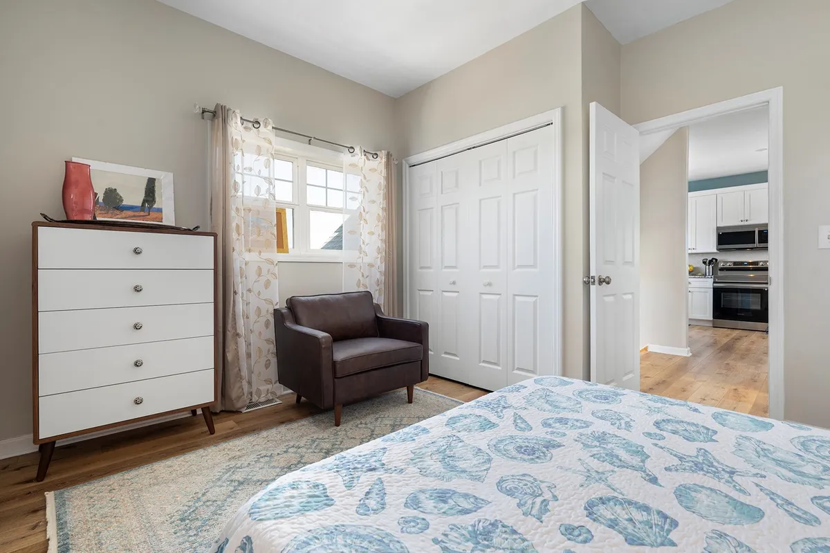 A cozy bedroom featuring a bed with a blue and white patterned quilt, a comfortable brown armchair, and a white dresser. Natural light streams in, creating a serene and inviting atmosphere ideal for relaxation and personal touches like bringing your own linens.